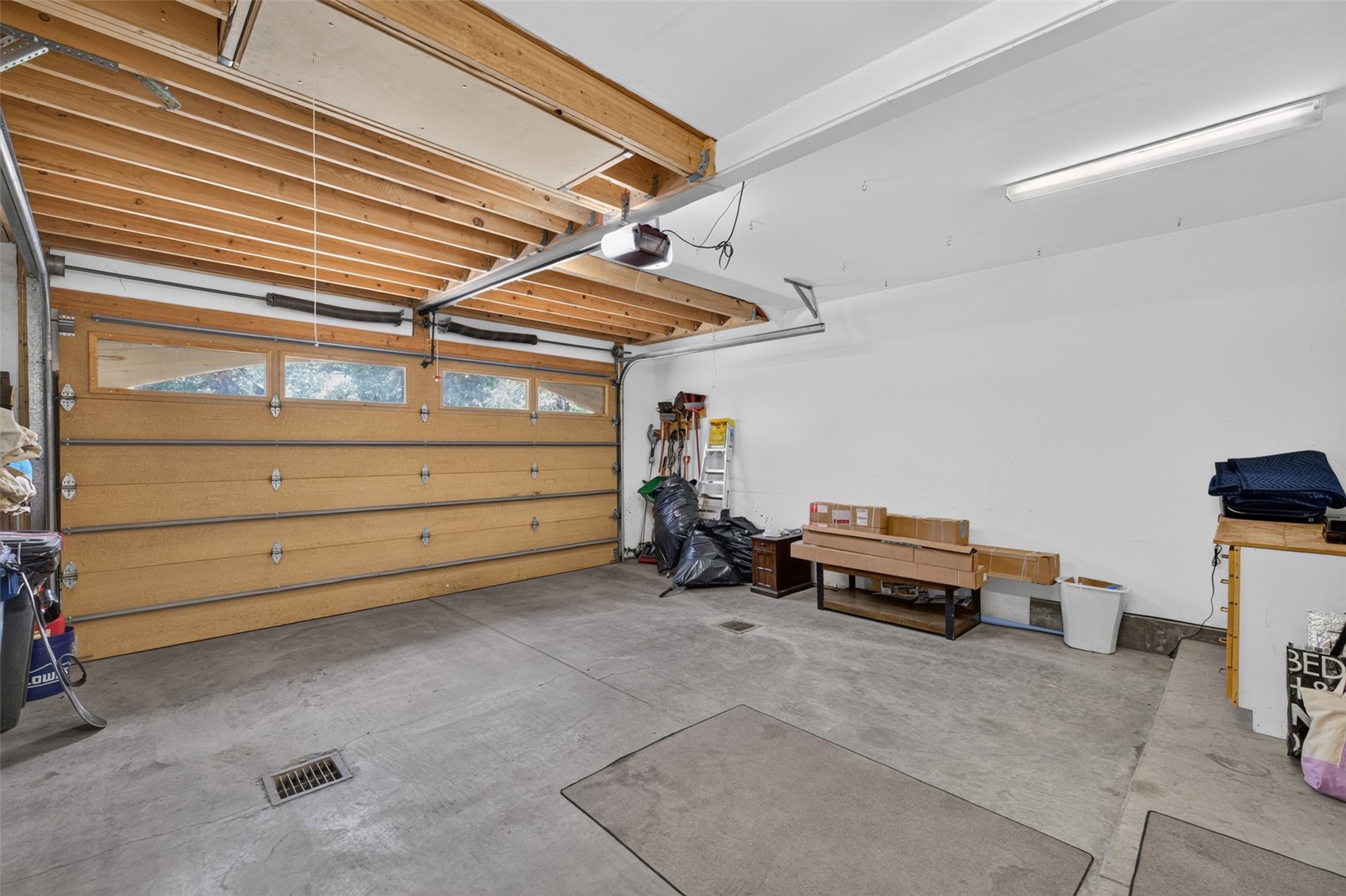 This image shows the interior of a spacious, unfinished garage with exposed wooden ceiling joists and a large, multi-paneled wooden garage door. The concrete floor features a central drainage grate, and the walls are painted white, providing a clean, utilitarian backdrop. Various storage items, including boxes, a ladder, and trash bags, are neatly tucked against the side wall, suggesting a functional workspace or storage area.