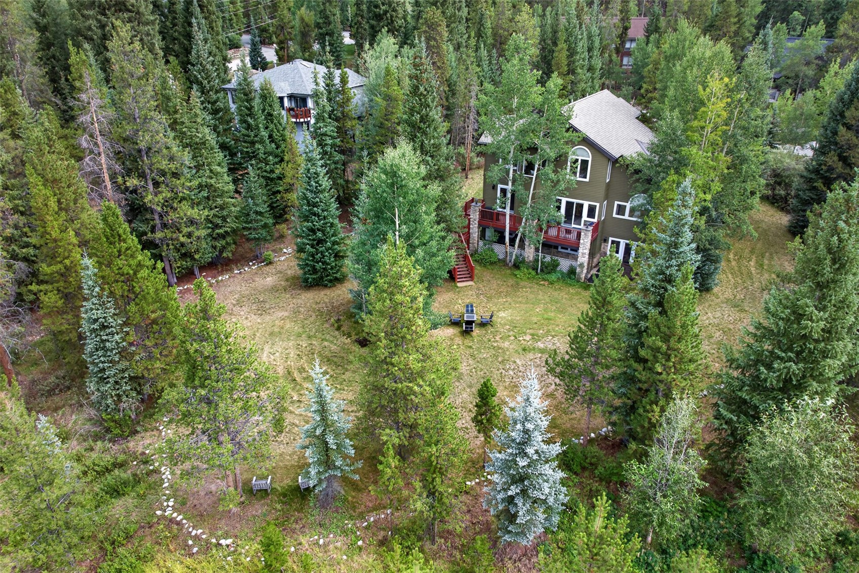 This high-angle aerial perspective captures a multi-story home nestled within a dense, lush forest of evergreen and deciduous trees. The property features a large, open grassy yard with a small seating area, a red-railed deck, and a stone-accented foundation. The setting offers a serene, secluded atmosphere, highlighting the home's integration into its natural, wooded environment.