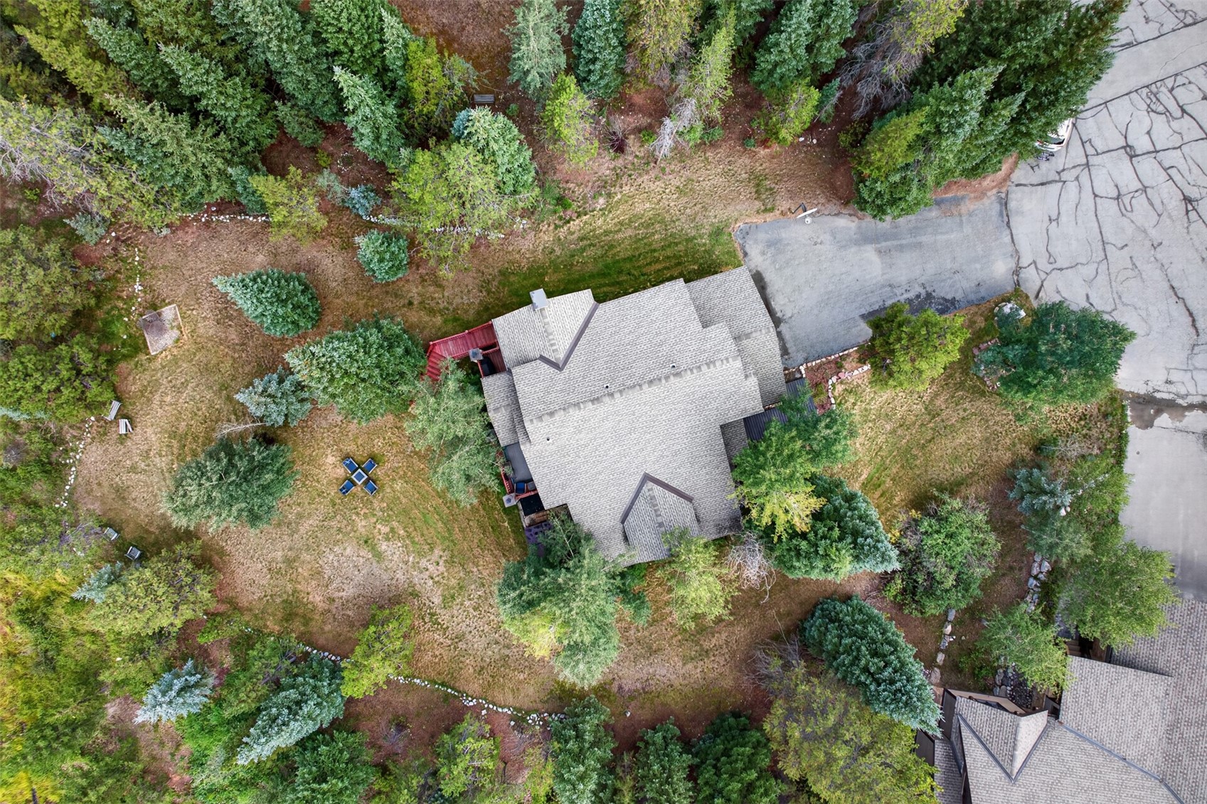 This high-angle aerial view captures a secluded residential property nestled within a dense forest of evergreen trees. The home features a complex, multi-gabled roofline and is surrounded by a mix of grassy lawn and natural woodland, with a paved driveway leading to the structure. A small outdoor seating area is visible on the lawn, emphasizing the quiet, private nature of the setting.