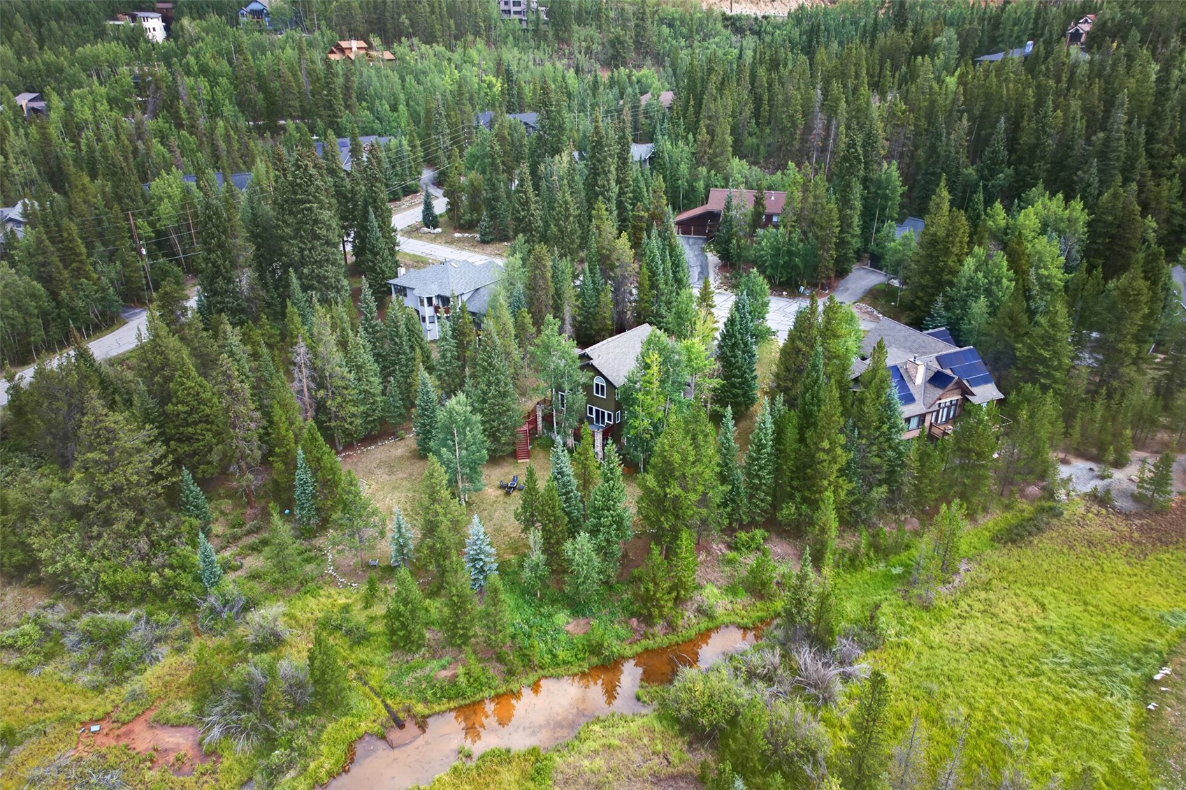 This high-angle aerial view captures a secluded mountain property nestled within a dense forest of evergreen and deciduous trees. The scene features several multi-story homes with pitched roofs, including one with prominent solar panels, set against a backdrop of lush, rolling terrain and a small, winding creek in the foreground. The perspective emphasizes the peaceful, natural setting and the integration of the architecture into the surrounding woodland landscape.