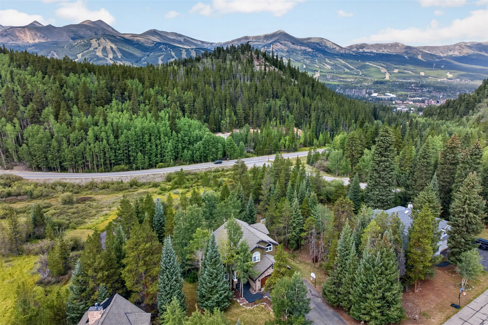 This high-angle aerial view captures a multi-story mountain home nestled within a dense forest of evergreen and deciduous trees. The property features a dark shingled roof, a paved driveway, and a scenic backdrop of expansive mountain ranges and a distant town. The perspective emphasizes the home's secluded, natural setting and its proximity to a winding mountain road.