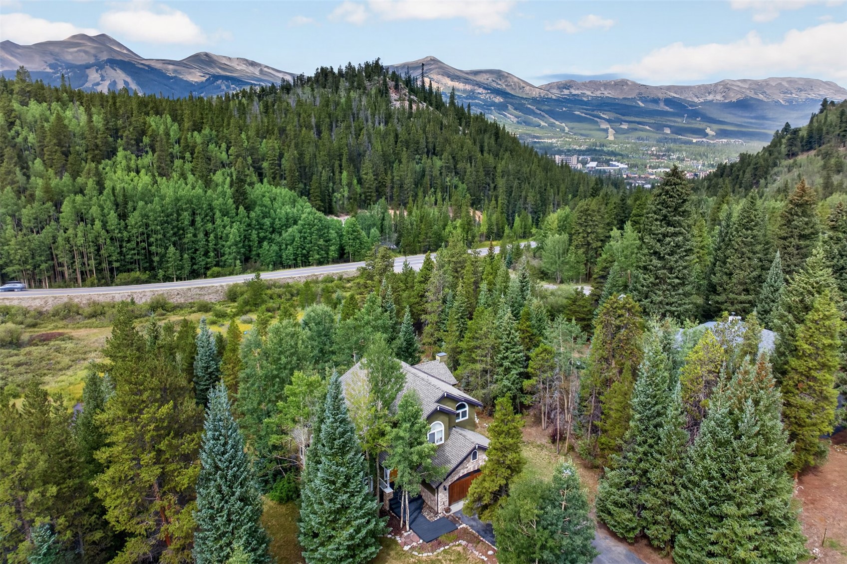 This high-angle aerial view captures a multi-story mountain home nestled within a dense forest of evergreen and deciduous trees. The property features a classic mountain-style architecture with a pitched roof, stone accents, and a paved driveway, set against a backdrop of expansive, rugged mountain ranges. The surrounding landscape is lush and verdant, emphasizing the home's secluded and scenic location.