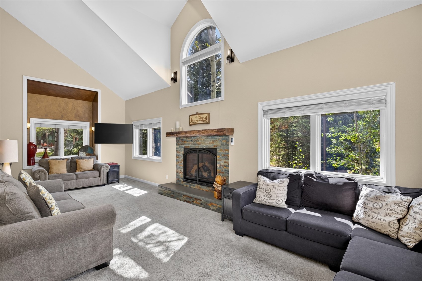 This spacious living room features a vaulted ceiling and a prominent stone-clad chimney breast centered between two large windows. The room is furnished with two plush sofas, one in a light gray and the other in a dark charcoal, arranged around a central seating area. A large arched window above the mantel allows natural light to flood the space, highlighting the neutral wall tones and carpeted flooring.