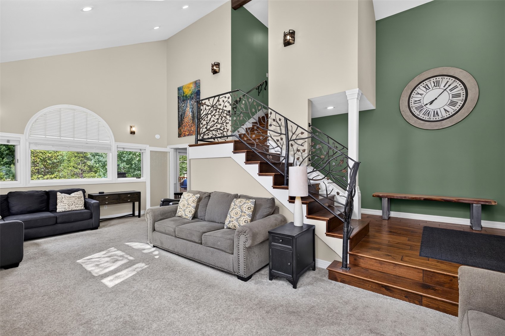 This spacious living area features a vaulted ceiling and a prominent staircase with decorative metal railings and dark wood treads. The room is anchored by a grey sofa and a black accent table, with a large arched window providing ample natural light. A striking green accent wall displays a large circular clock, while the overall aesthetic blends traditional architectural elements with contemporary furnishings.
