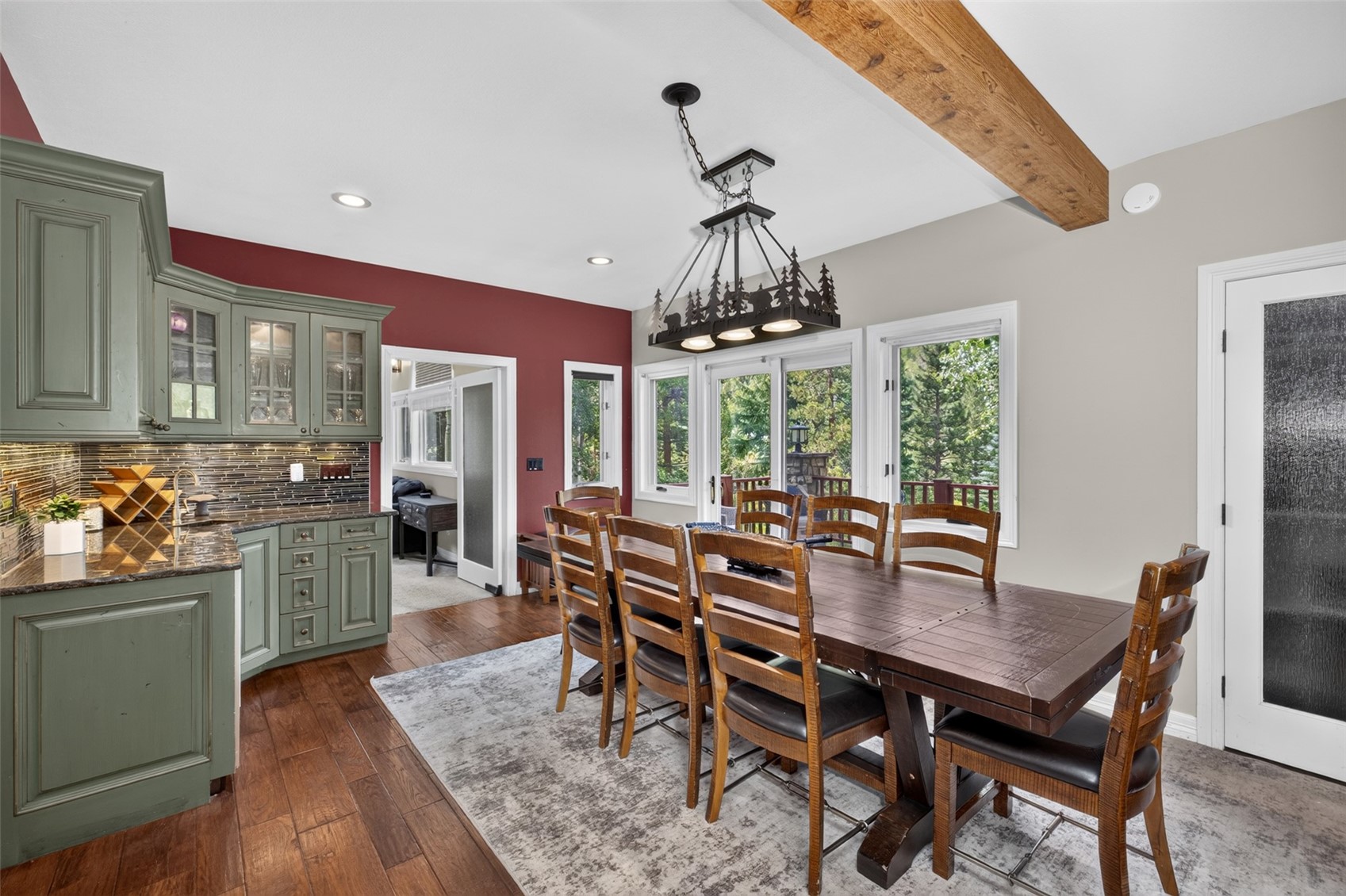 This dining room features a rustic aesthetic with a large wooden table surrounded by six chairs, positioned beneath a decorative chandelier with a forest-themed silhouette. The space is anchored by a patterned area rug on dark wood flooring and includes a built-in green cabinetry unit with a dark stone countertop. Large sliding glass doors provide natural light and a view of the surrounding trees, while a wooden ceiling beam adds architectural character to the room.
