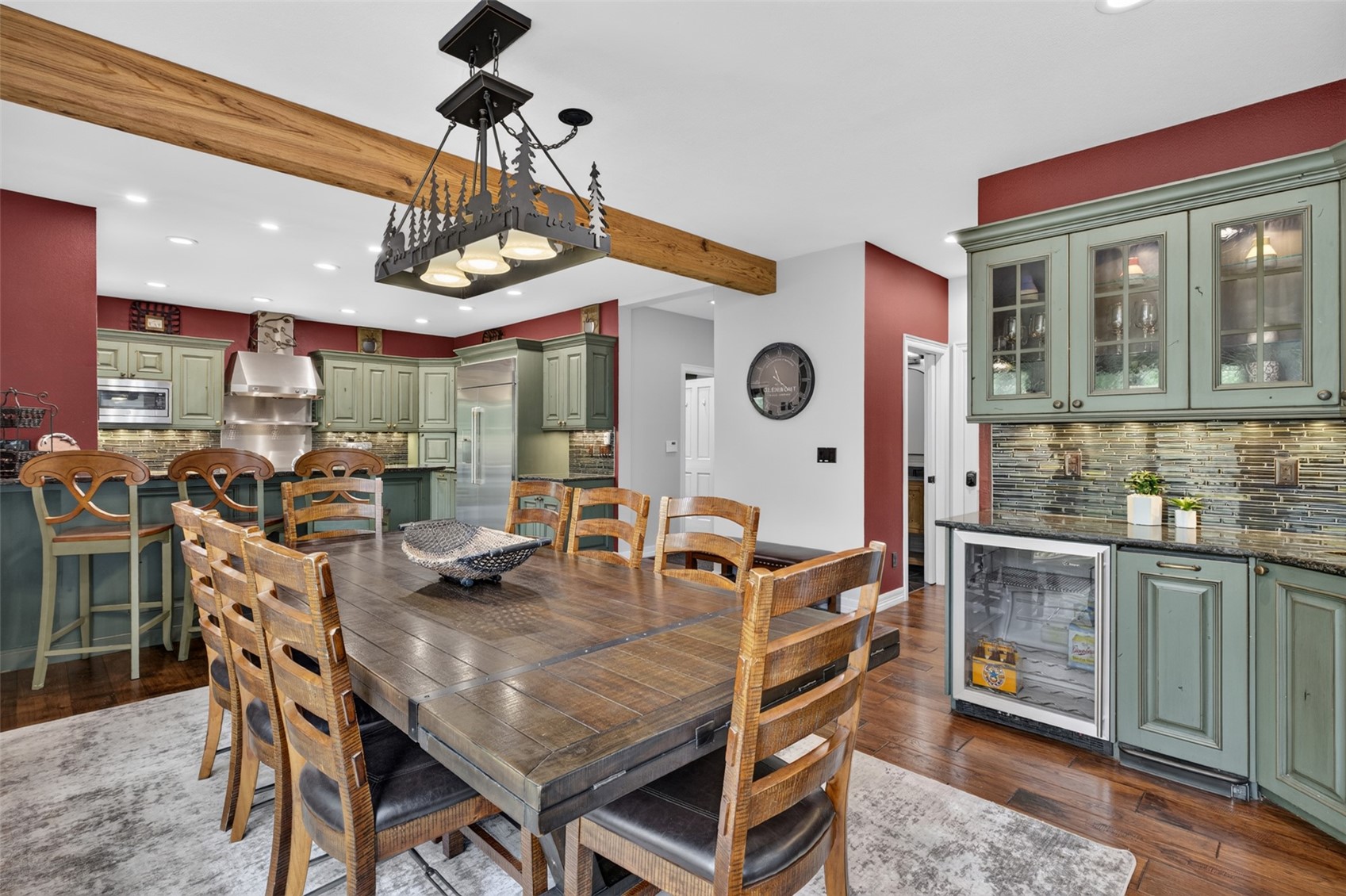 This open-concept kitchen and dining area features a rustic aesthetic with sage green cabinetry, a large wooden dining table, and a distinctive forest-themed chandelier. The space is anchored by a prominent wooden ceiling beam and deep red accent walls, creating a warm, lodge-inspired atmosphere. A built-in beverage refrigerator and glass-front display cabinets add functional elegance to the kitchen perimeter.