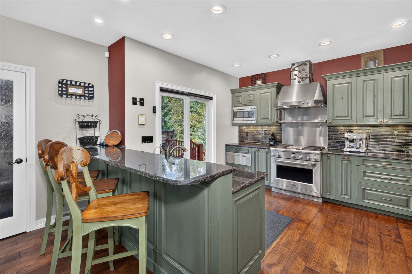 This kitchen features sage green cabinetry paired with dark stone countertops and a tiled backsplash, creating a warm and inviting atmosphere. A central island with a breakfast bar provides seating for three, while stainless steel appliances, including a professional-style range and hood, add a modern touch. The space is finished with rich wood flooring and recessed lighting, with a sliding glass door providing a view of the outdoor greenery.