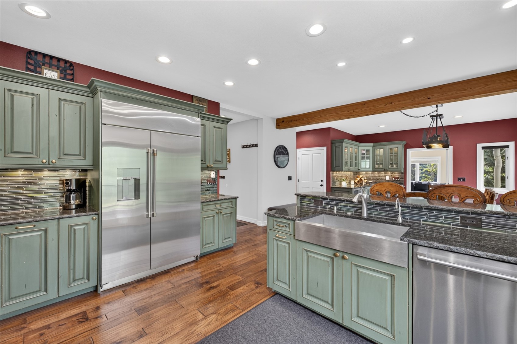 This spacious kitchen features distinctive sage green cabinetry with a distressed finish, paired with dark stone countertops and a tiled backsplash. A large stainless steel farmhouse sink is centered in the island, while a professional-grade refrigerator stands prominently against the wall. The room is accented by a rustic wooden ceiling beam and warm hardwood flooring, creating a cozy yet functional culinary space.
