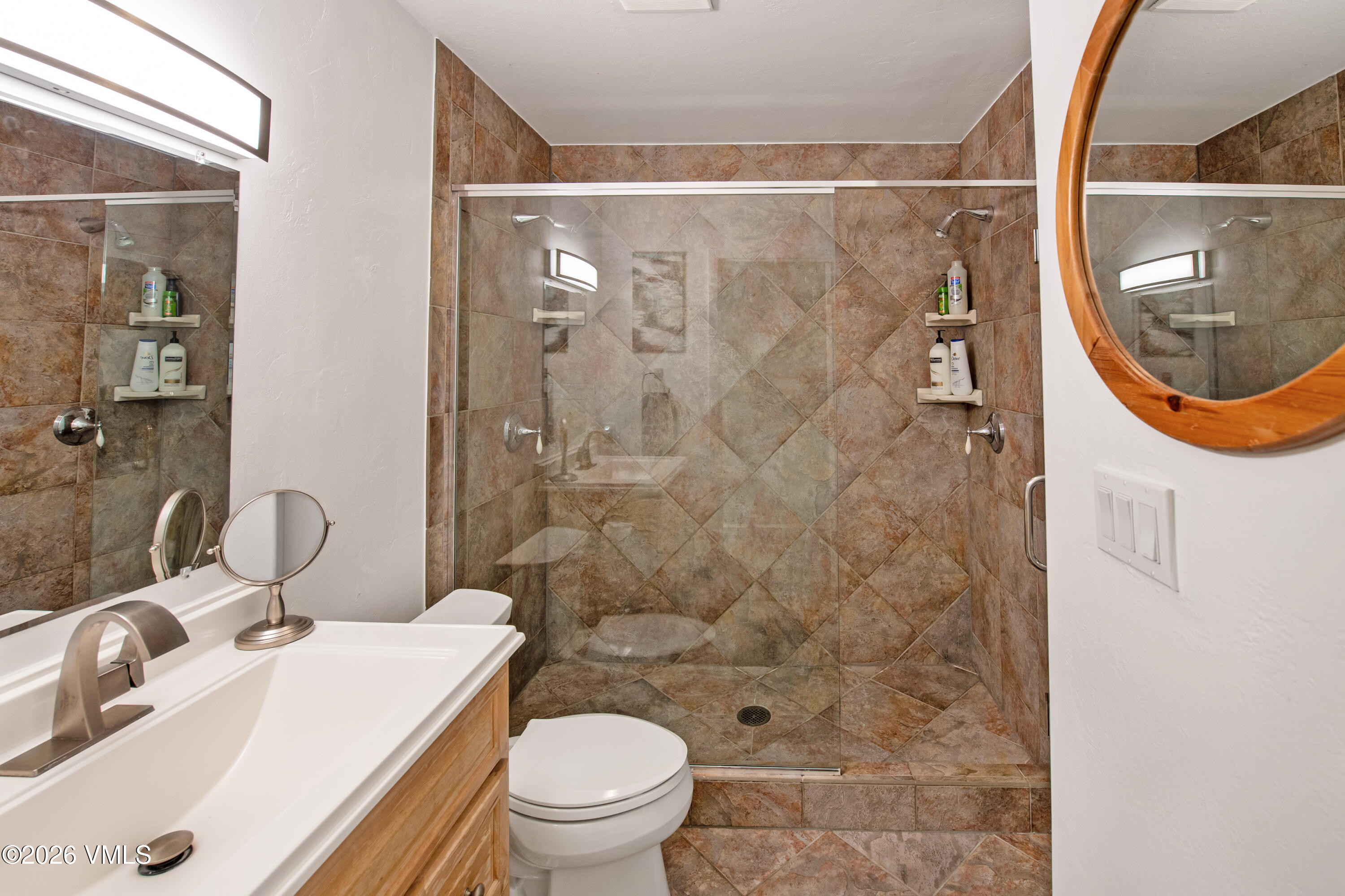 This compact bathroom features a walk-in shower with brown patterned tile walls and a glass door, adjacent to a white toilet. A light wood vanity with a white countertop and a modern faucet sits below a large mirror, while a round wall-mounted mirror adds a functional touch. The space is well-lit by a sleek vanity light fixture and a circular mirror on the opposite wall, creating a clean and practical layout.
