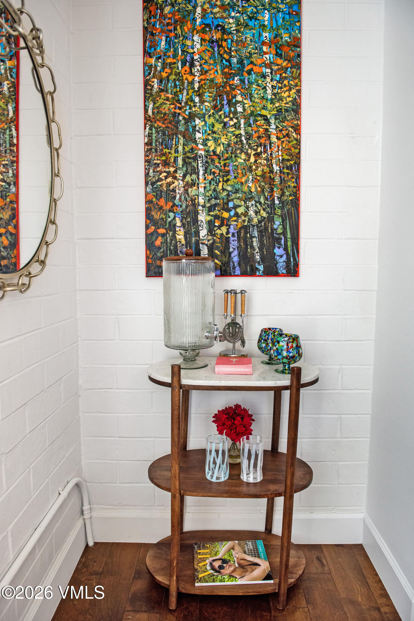 This image captures a decorative vignette featuring a three-tiered wooden stand with a stone top, positioned against a white painted brick wall. The stand holds a glass beverage dispenser, colorful glassware, a small vase of red flowers, and a magazine, while a vibrant abstract forest painting hangs above it. To the left, a large oval mirror with a decorative chain frame adds a touch of elegance to this curated interior display.
