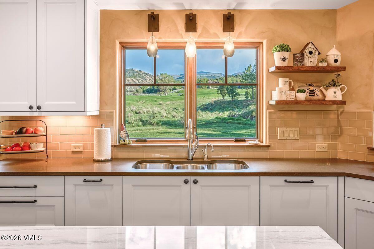 This bright and inviting kitchen features crisp white cabinetry paired with a warm, neutral-toned backsplash and dark countertops. A central window provides a picturesque view of a lush green landscape, while open shelving to the right displays charming decorative accents. The space is illuminated by three pendant lights, creating a cozy and functional atmosphere.