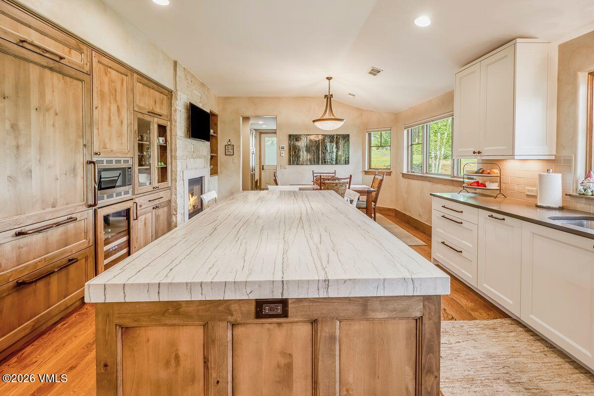 This spacious kitchen features a large central island with a light-toned, veined stone countertop that serves as the focal point of the room. The space combines warm wood cabinetry on the left with crisp white cabinets on the right, creating a balanced, transitional aesthetic. A stone-clad chimney breast with an integrated fireplace adds a cozy element, while the open layout flows seamlessly into a dining area bathed in natural light from multiple windows.