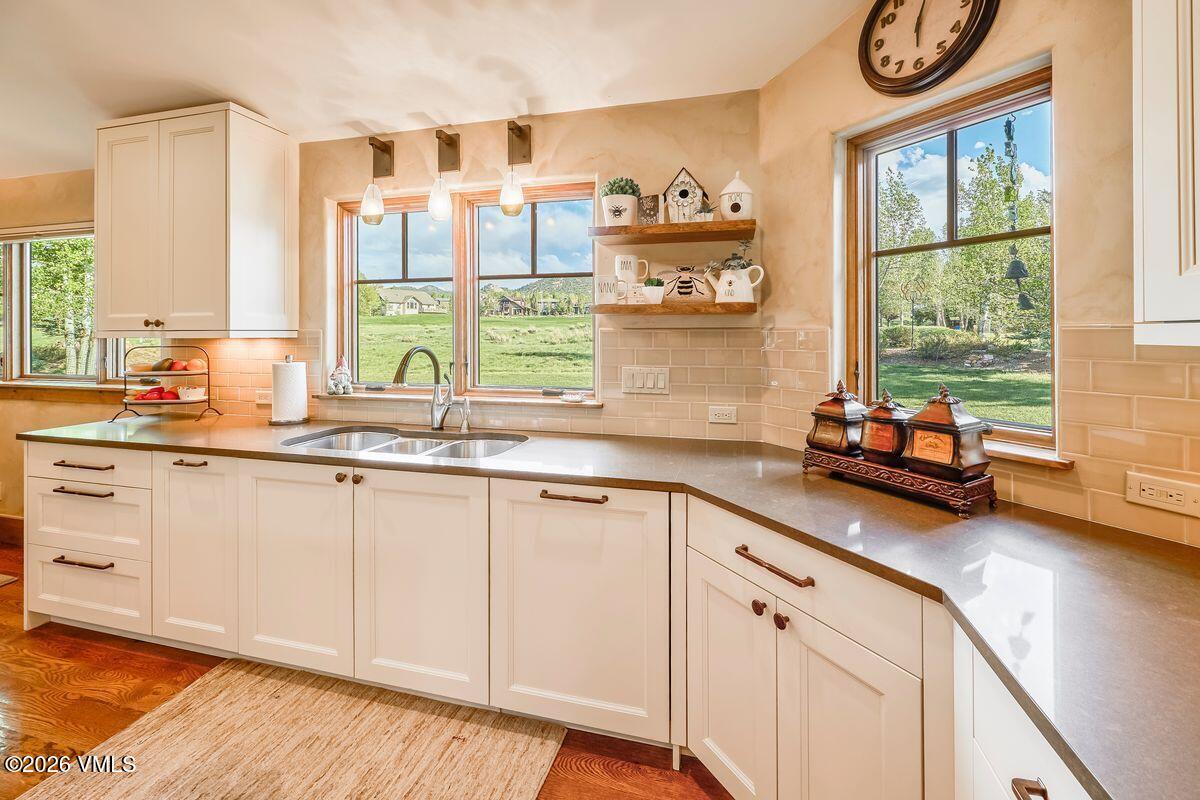 This bright and inviting kitchen features white cabinetry paired with neutral-toned countertops and a matching subway tile backsplash. A double-basin sink is positioned beneath two windows that offer a scenic view of the surrounding landscape, while open shelving provides a charming display area for decor. The space is well-lit by natural light and pendant fixtures, creating a warm and functional atmosphere.