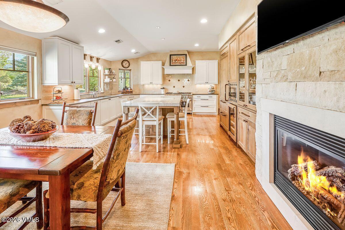 This open-concept kitchen and dining area features a warm, rustic aesthetic with light wood flooring and white cabinetry. A central island with bar seating serves as a focal point, while a stone-clad chimney breast housing a fireplace adds a cozy element to the space. The perspective captures the flow from the dining table in the foreground through the kitchen to the rear windows, creating an inviting and spacious atmosphere.