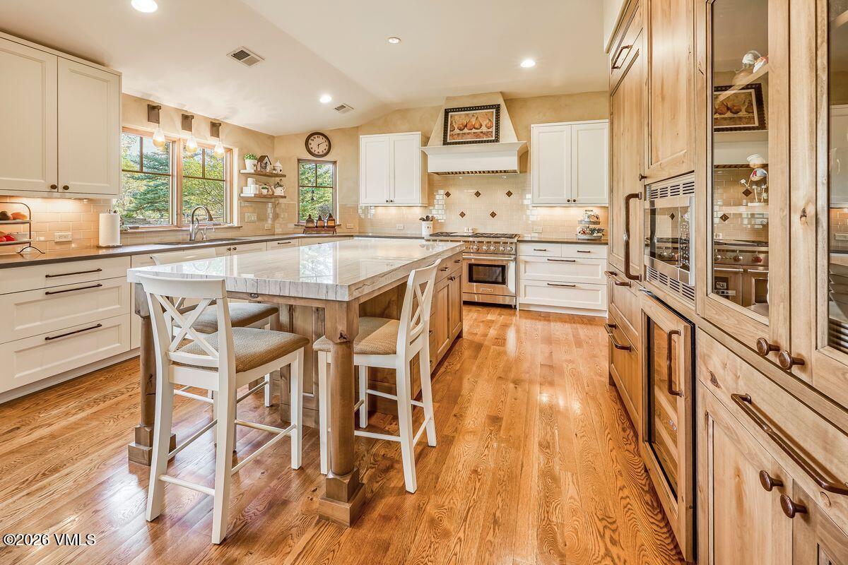 This spacious kitchen features a large central island with a stone countertop and seating for two, complemented by white cabinetry and warm wood-toned accents. The room is illuminated by recessed lighting and natural light from the windows above the sink, creating an inviting and functional atmosphere. A professional-style range with a decorative hood serves as a focal point, while the warm wood flooring adds a cohesive, traditional feel to the space.