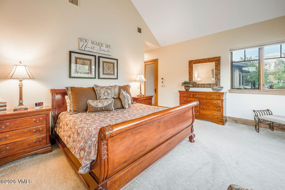 This spacious primary bedroom features a large wooden sleigh bed as the focal point, complemented by matching nightstands and a dresser topped with a decorative mirror. The room is bathed in natural light from a set of windows on the right, while a vaulted ceiling adds a sense of openness to the space. Neutral wall tones and soft carpeting create a warm, inviting atmosphere, accented by framed wall art above the bed.