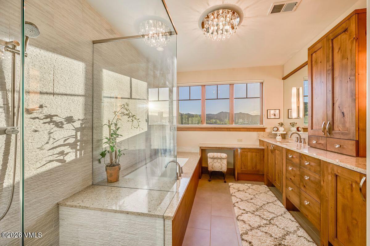 This primary bathroom features a luxurious soaking tub enclosed by a glass partition, set against a textured stone wall. A long vanity with warm-toned wood cabinetry and a stone countertop provides ample storage, while a dedicated makeup vanity area sits beneath a large window overlooking the landscape. The space is illuminated by a decorative crystal chandelier, creating an elegant and serene atmosphere.