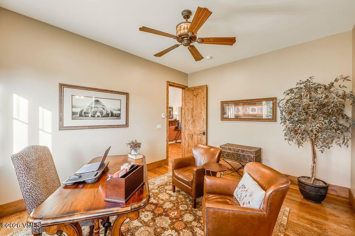 This well-appointed home office features a rich wood desk, two comfortable leather armchairs, and a patterned area rug that anchors the space. A ceiling fan with wooden blades hangs above, while a large framed bear print and a decorative wall art piece add character to the neutral-toned walls. The room is brightened by natural light from a window on the left, and a doorway leads into an adjacent room, creating an inviting and professional atmosphere.