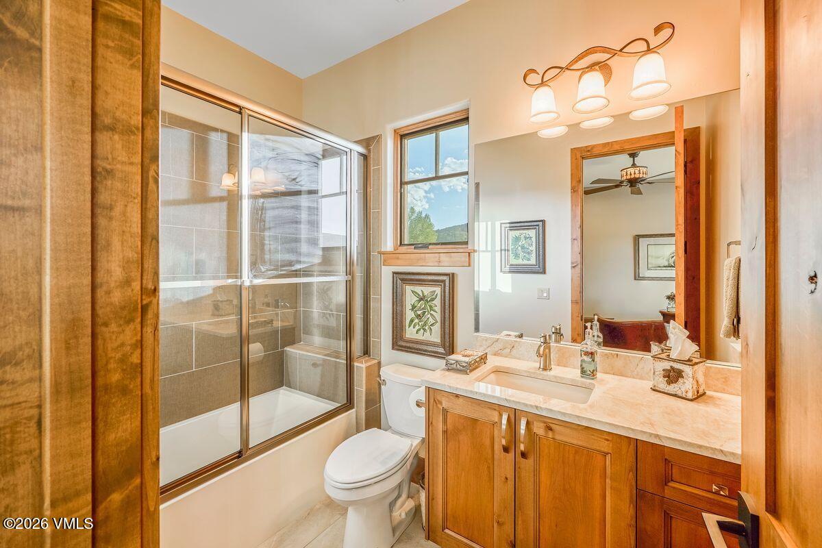 This compact bathroom features warm wood cabinetry topped with a light-toned stone vanity and a rectangular undermount sink. A glass-enclosed shower with neutral-toned wall tiles sits adjacent to the toilet, while a large mirror above the vanity reflects the room and a window. The space is illuminated by a three-light fixture and natural light from the window, creating a cozy and functional atmosphere.