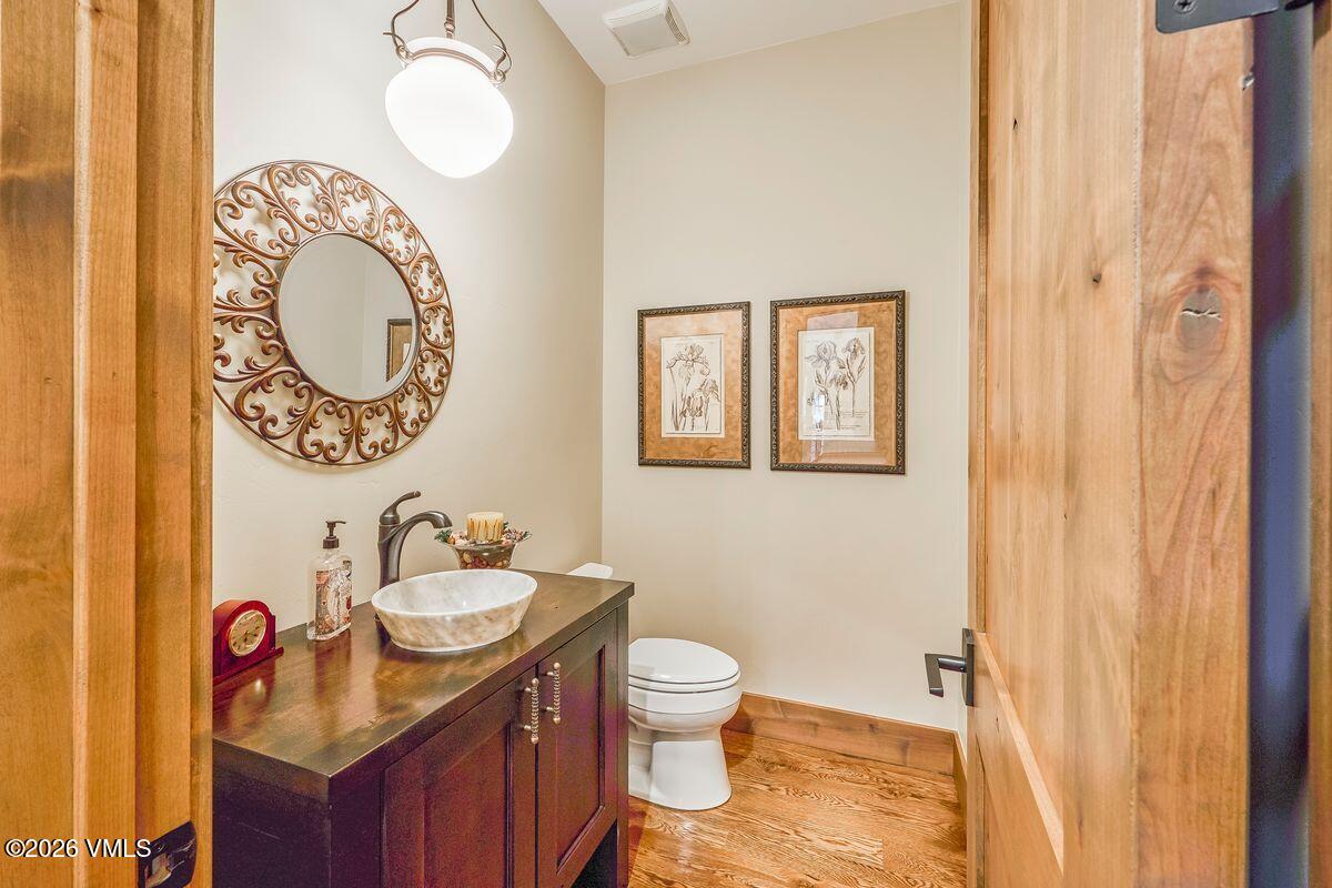 This compact powder room features a rustic aesthetic with a dark wood vanity topped by a stone vessel sink and a decorative, ornate circular mirror. The walls are painted a neutral tone, complemented by two framed botanical prints and warm wood flooring. The perspective is framed through a wooden doorway, highlighting the cozy and well-appointed nature of the space.