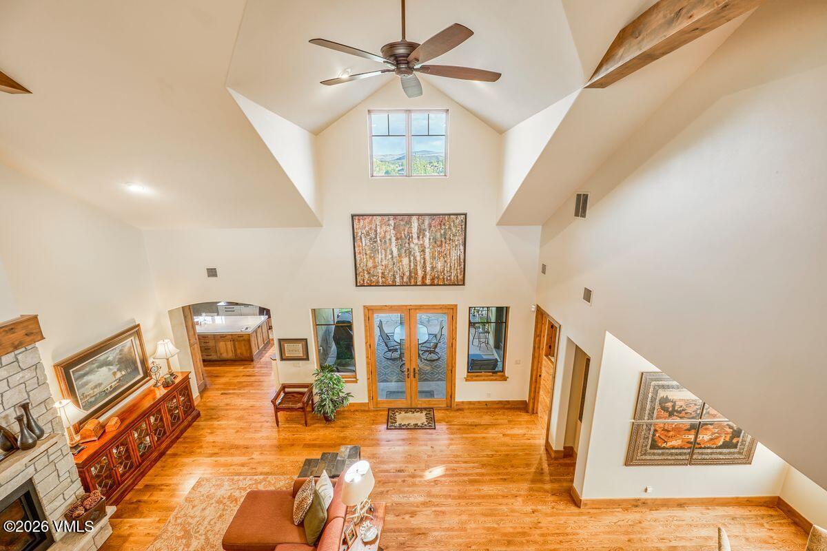 This high-angle view captures a spacious, open-concept living area characterized by a dramatic vaulted ceiling and warm wood flooring. A stone-clad chimney breast anchors the left side of the room, while a large piece of abstract art serves as a focal point on the central wall above a set of glass-paned double doors. The space is bathed in natural light from multiple windows, creating an airy and inviting atmosphere that flows seamlessly into an adjacent kitchen area.