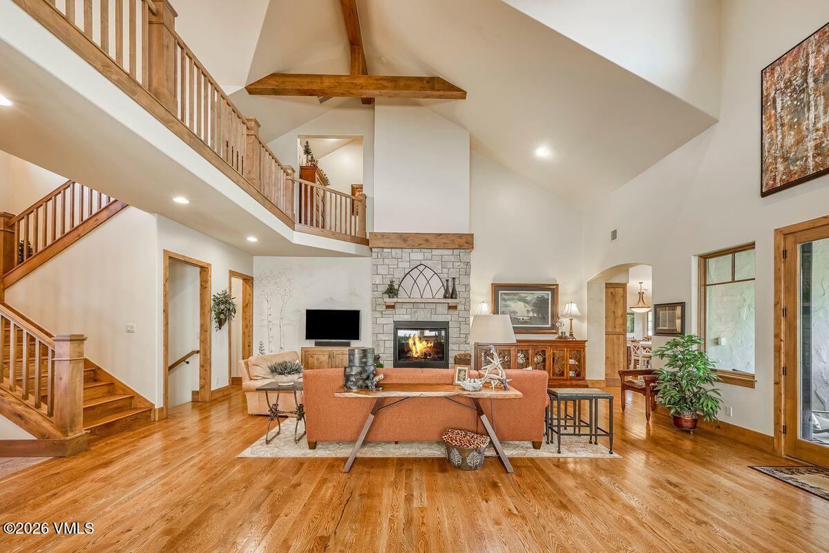 This spacious living room features a vaulted ceiling with exposed wooden beams and a prominent stone-clad chimney breast housing a fireplace. The room is anchored by a warm-toned sofa and a wooden console table, with light wood flooring extending throughout the open-concept space. A staircase with wooden railings leads to an upper-level landing, creating an airy, multi-level perspective that emphasizes the home's open architecture.