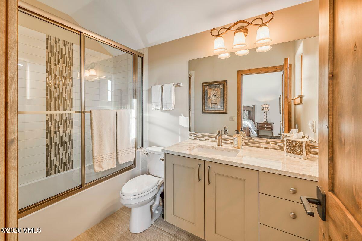 This well-appointed bathroom features a neutral color palette with light-toned cabinetry and a stone-topped vanity. A glass-enclosed shower with decorative tile accents sits beside a standard toilet, while a large mirror reflects the adjacent bedroom space. The room is illuminated by a decorative three-light fixture, creating a bright and functional atmosphere.