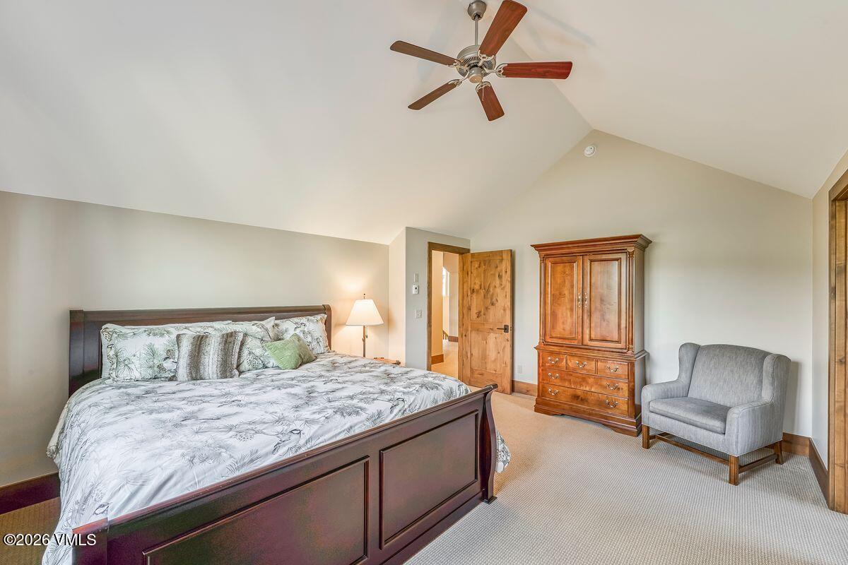 This spacious primary bedroom features a vaulted ceiling with a central wooden ceiling fan and neutral-toned walls. The room is anchored by a large dark wood bed frame with a patterned duvet, complemented by a matching wooden armoire and a comfortable gray armchair in the corner. The space is carpeted throughout and includes a doorway leading to an adjacent hallway, creating an open and inviting atmosphere.
