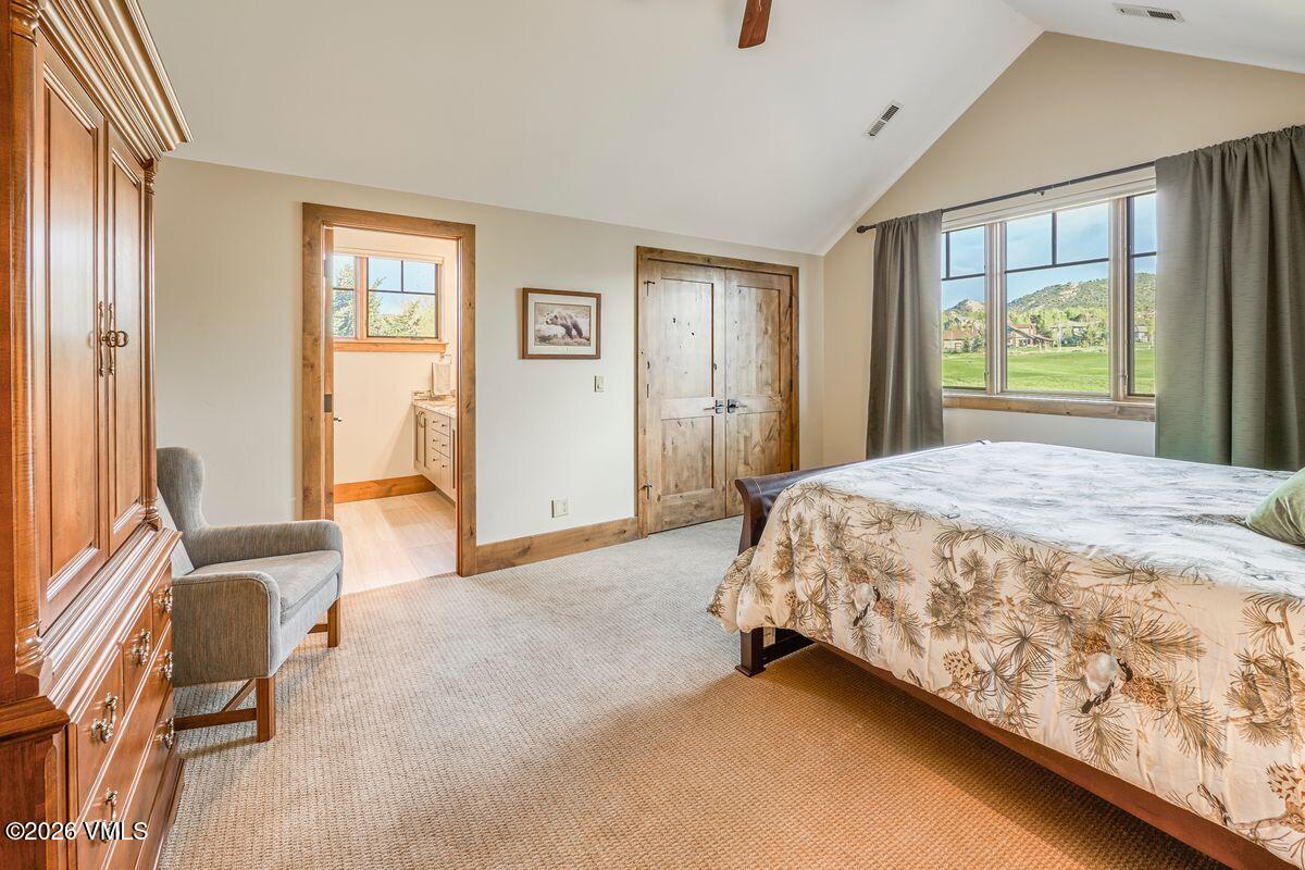 This spacious bedroom features a vaulted ceiling and a large window that offers a scenic view of the surrounding landscape. The room is furnished with a bed featuring a patterned quilt, a comfortable armchair, and a large wooden armoire, all set against neutral-toned walls. A doorway leads into an adjacent bathroom, and the space is accented by rustic wooden doors and trim, creating a warm and inviting atmosphere.