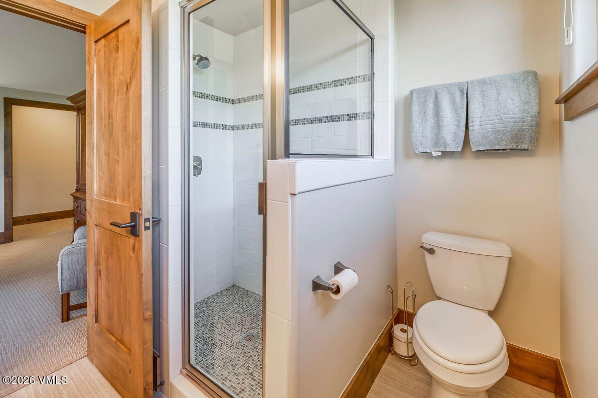 This compact bathroom features a glass-enclosed shower with a pebble-style floor and white wall tiles. A standard white toilet is positioned next to the shower, with a wall-mounted toilet paper holder and a towel rack holding two grey towels. The space is finished with light-toned wood trim and neutral wall paint, creating a clean and functional aesthetic.