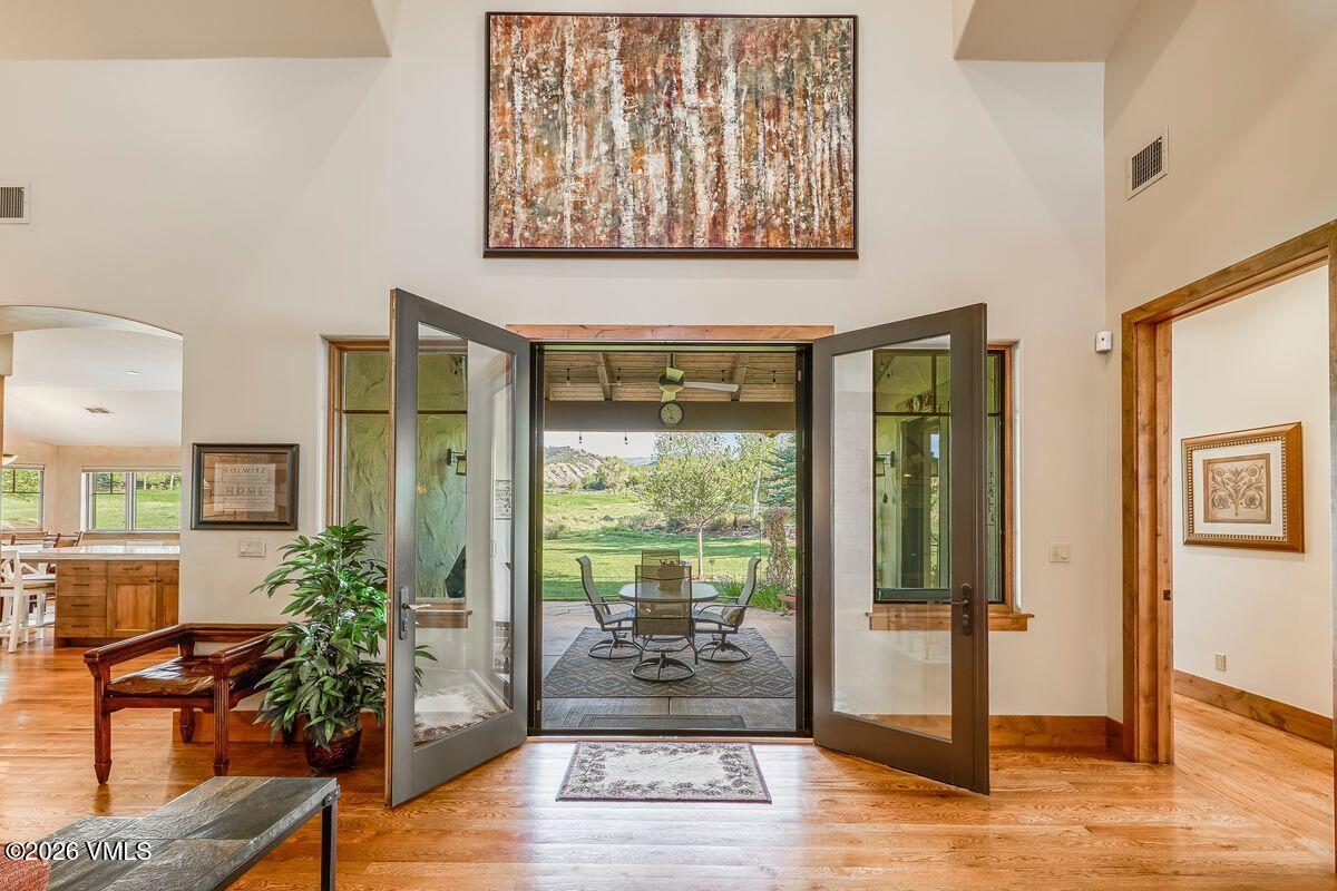 This interior view captures a bright living space featuring warm wood flooring and a large, abstract painting mounted high on the wall. Double glass doors stand open, creating a seamless transition to a covered outdoor patio area furnished with a dining set. The perspective looks directly through the entryway, highlighting the connection between the indoor living area and the expansive, scenic backyard landscape.