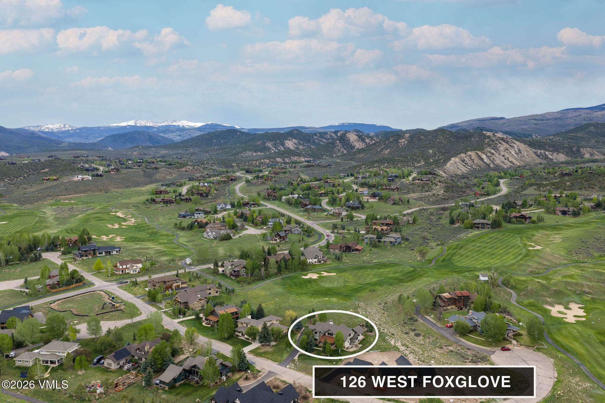 This high-altitude aerial shot captures a sprawling residential community nestled within a lush, green valley surrounded by distant, snow-capped mountains. The property at 126 West Foxglove is highlighted within a white oval, showcasing its position adjacent to a manicured golf course and winding neighborhood roads. The perspective provides a comprehensive view of the landscape, emphasizing the scenic, quiet nature of the location.
