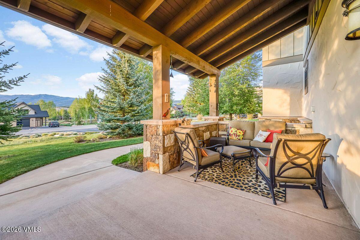 This inviting covered patio features a rustic timber-framed structure with a stone-clad pillar, creating a cozy outdoor living space. The area is furnished with comfortable cushioned seating arranged on a patterned rug, overlooking a lush green lawn and mature pine trees. The perspective captures the transition from the shaded porch to the bright, open landscape, emphasizing a peaceful and well-maintained residential setting.