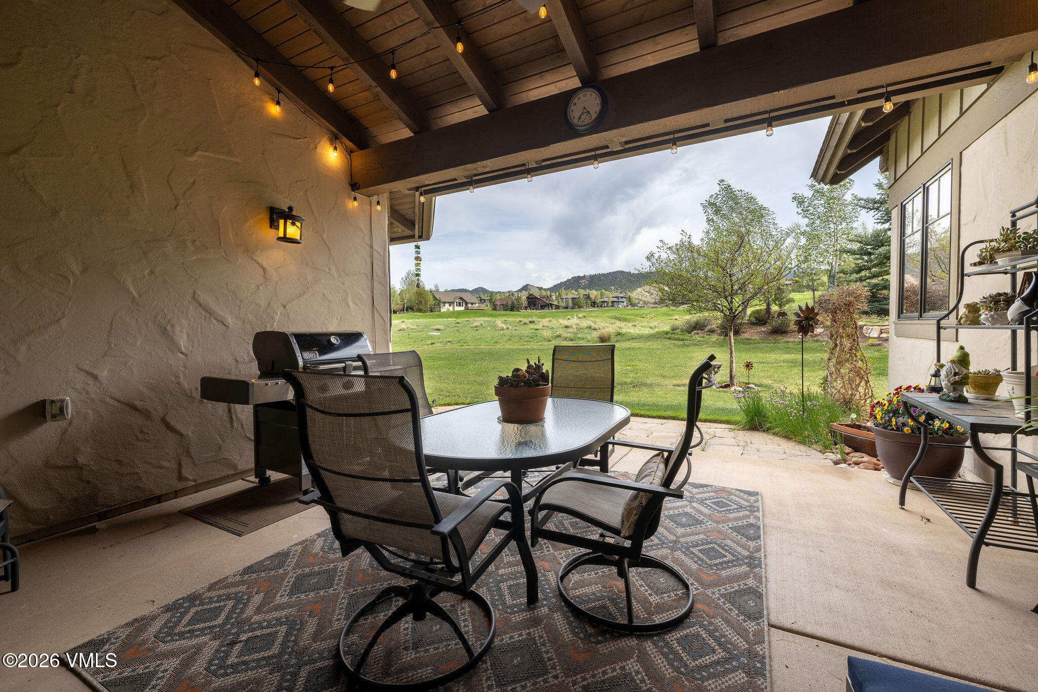 This covered patio offers a comfortable outdoor living space featuring a glass-topped dining set, a grill, and a patterned area rug. The space is framed by a rustic wood-beamed ceiling with string lights, creating an inviting atmosphere that opens directly onto a lush green lawn with mountain views. The perspective is from under the covered area, looking out toward the expansive backyard and surrounding landscape.