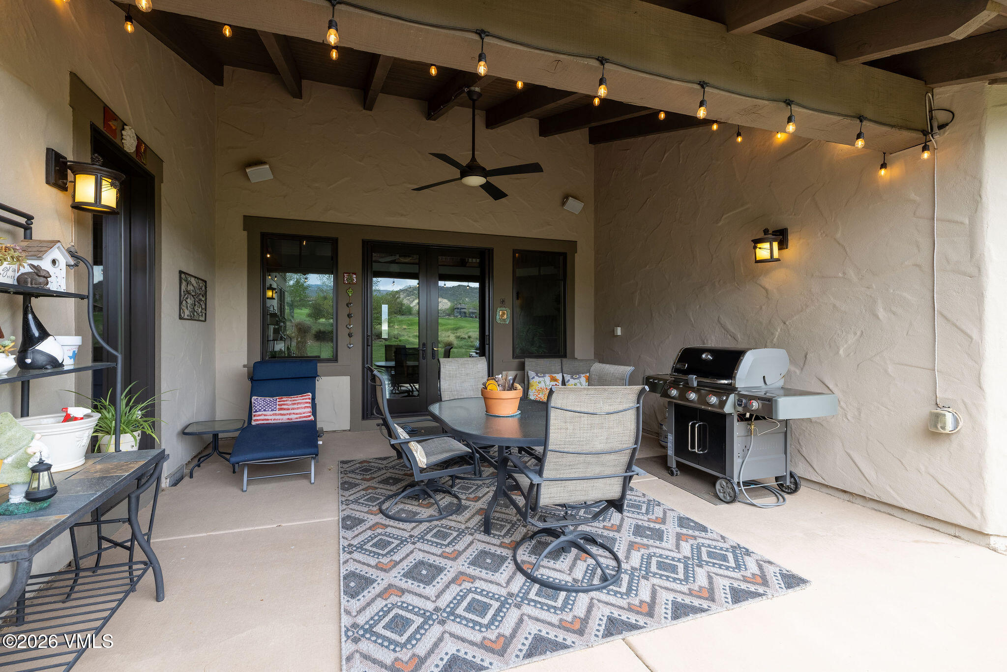 This covered patio space offers a comfortable outdoor living area featuring a dining set with four chairs, a lounge chair with a patriotic-themed cushion, and a stainless steel grill. The space is illuminated by string lights and wall-mounted fixtures, with a ceiling fan providing airflow under the exposed beam ceiling. Large glass doors provide a clear view of the surrounding landscape, creating a seamless transition between the indoor and outdoor environments.
