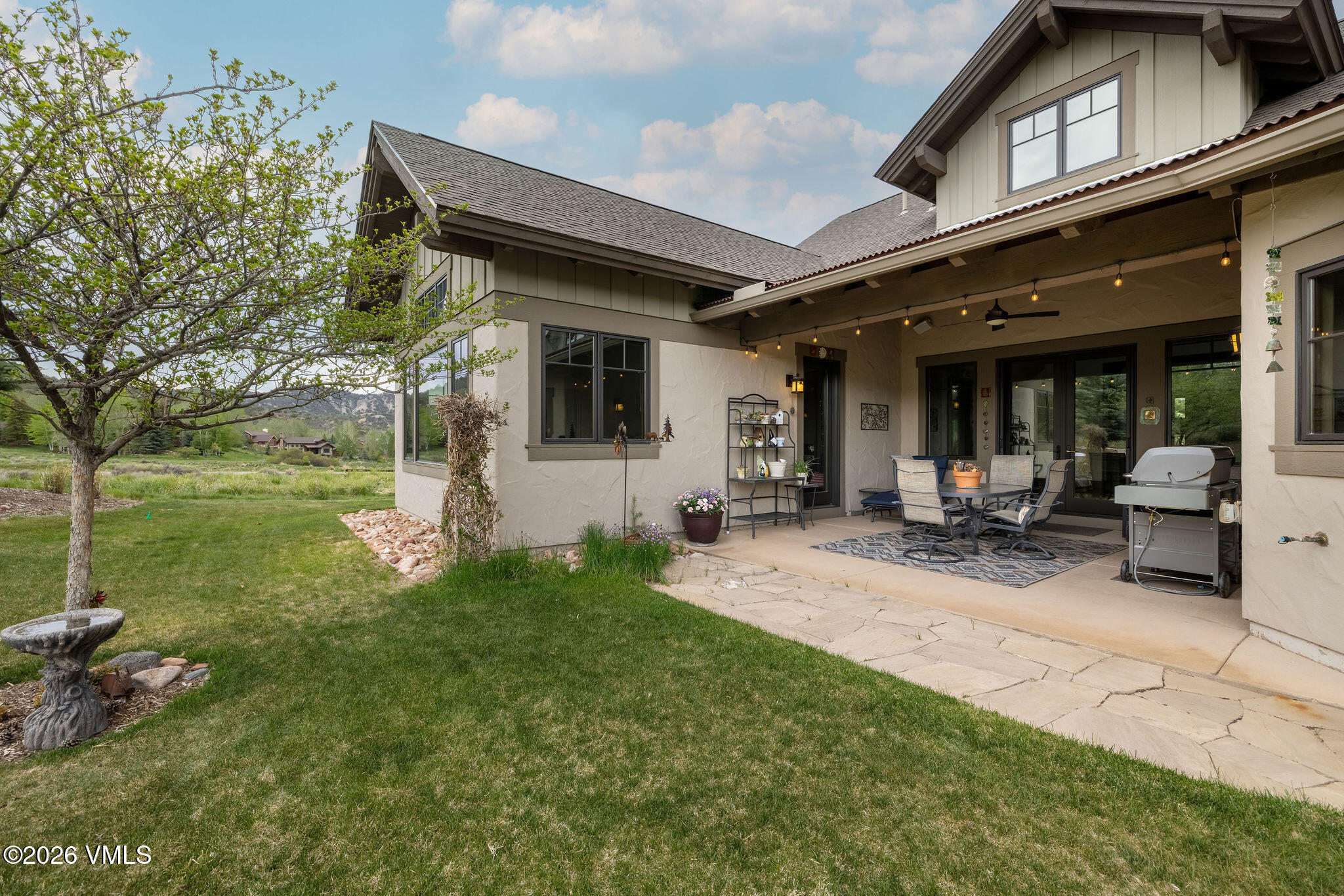 This image captures a spacious covered patio area attached to a home with a neutral-toned stucco exterior and dark trim. The patio features a stone-paved floor, a dining set, and a grill, creating an inviting outdoor living space that overlooks a lush green lawn and a scenic landscape. The perspective is from the yard, looking toward the house, highlighting the seamless transition between the indoor living area and the natural surroundings.