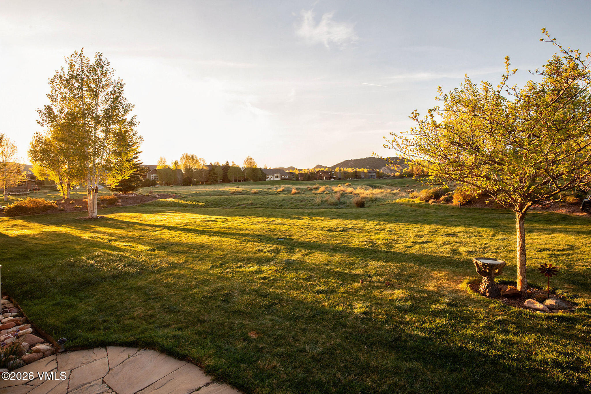This expansive backyard features a lush, well-maintained lawn that transitions into a natural, open meadow area. The scene is framed by mature trees on either side, with a stone birdbath positioned near a tree in the foreground and a stone patio edge visible at the bottom. The warm, golden light of late afternoon casts long shadows across the grass, creating a serene and inviting atmosphere with distant mountain views.