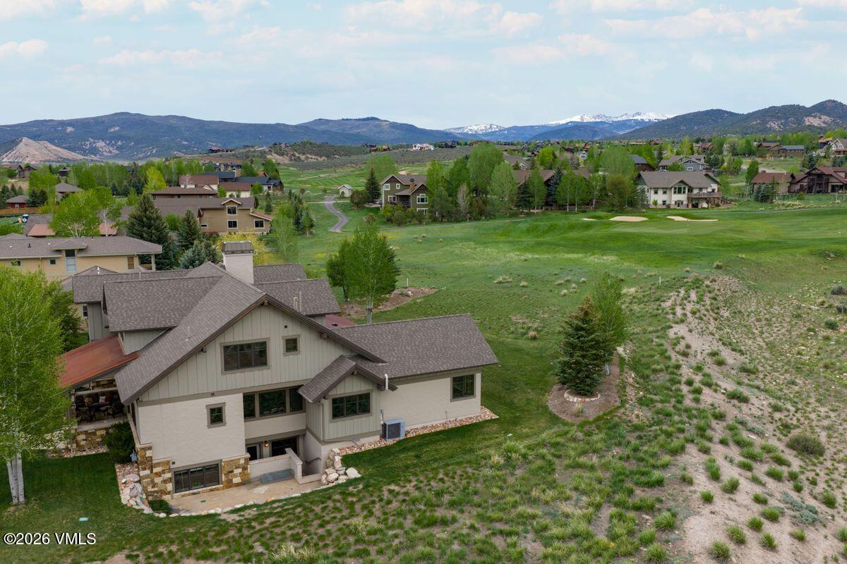 This elevated aerial perspective captures a large, multi-level mountain-style residence nestled against a sprawling green golf course. The home features a complex roofline with multiple gables, a mix of light-colored stucco and wood siding, and a stone-accented foundation. Beyond the property, the landscape opens up to rolling hills and distant snow-capped mountains under a bright, partly cloudy sky.