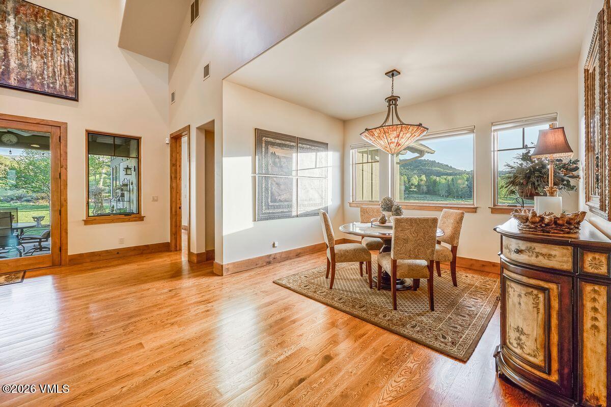 This elegant dining area features warm wood flooring and a central table surrounded by four upholstered chairs, all set atop a patterned area rug. A decorative pendant light hangs above the table, while large windows provide ample natural light and views of the surrounding landscape. To the right, a detailed wooden cabinet with a lamp adds a touch of traditional charm to the space.