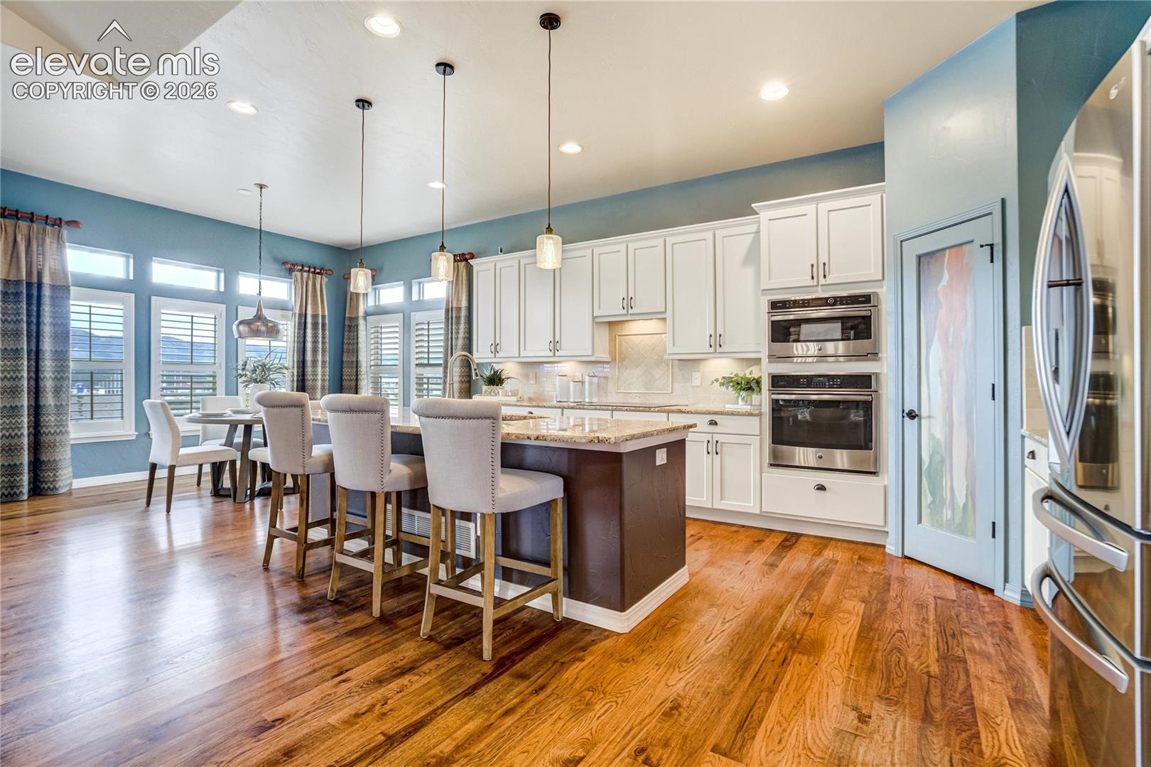 This bright and open-concept kitchen features white cabinetry paired with a dark-toned kitchen island and warm-toned wood flooring. The space is illuminated by three pendant lights hanging over the island and recessed ceiling lighting, while a dining area with a round table sits adjacent to large windows. The kitchen is equipped with stainless steel appliances, including a built-in oven and microwave, and features a pantry door with decorative glass.