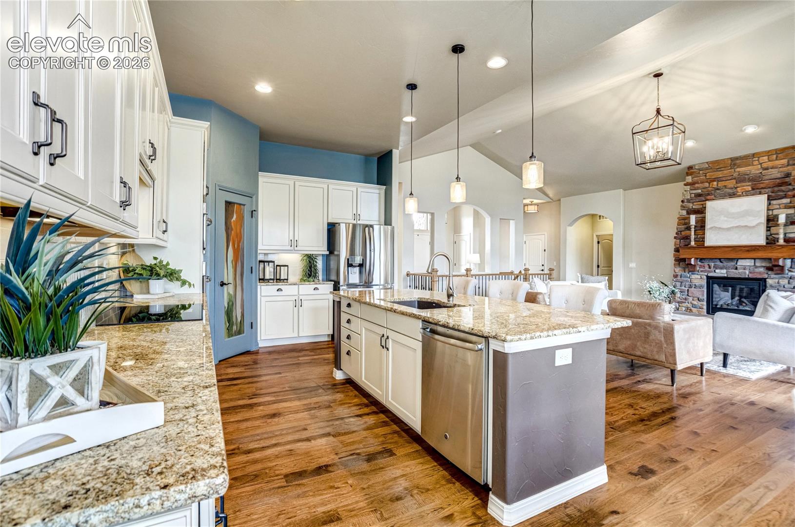 This open-concept kitchen features white cabinetry, granite countertops, and a central island with a stainless steel dishwasher and sink. The space flows seamlessly into a living area anchored by a stone-clad chimney breast and a wooden mantel. Warm wood flooring runs throughout the home, complemented by pendant lighting and a blue-accented pantry door, creating a bright and inviting atmosphere.