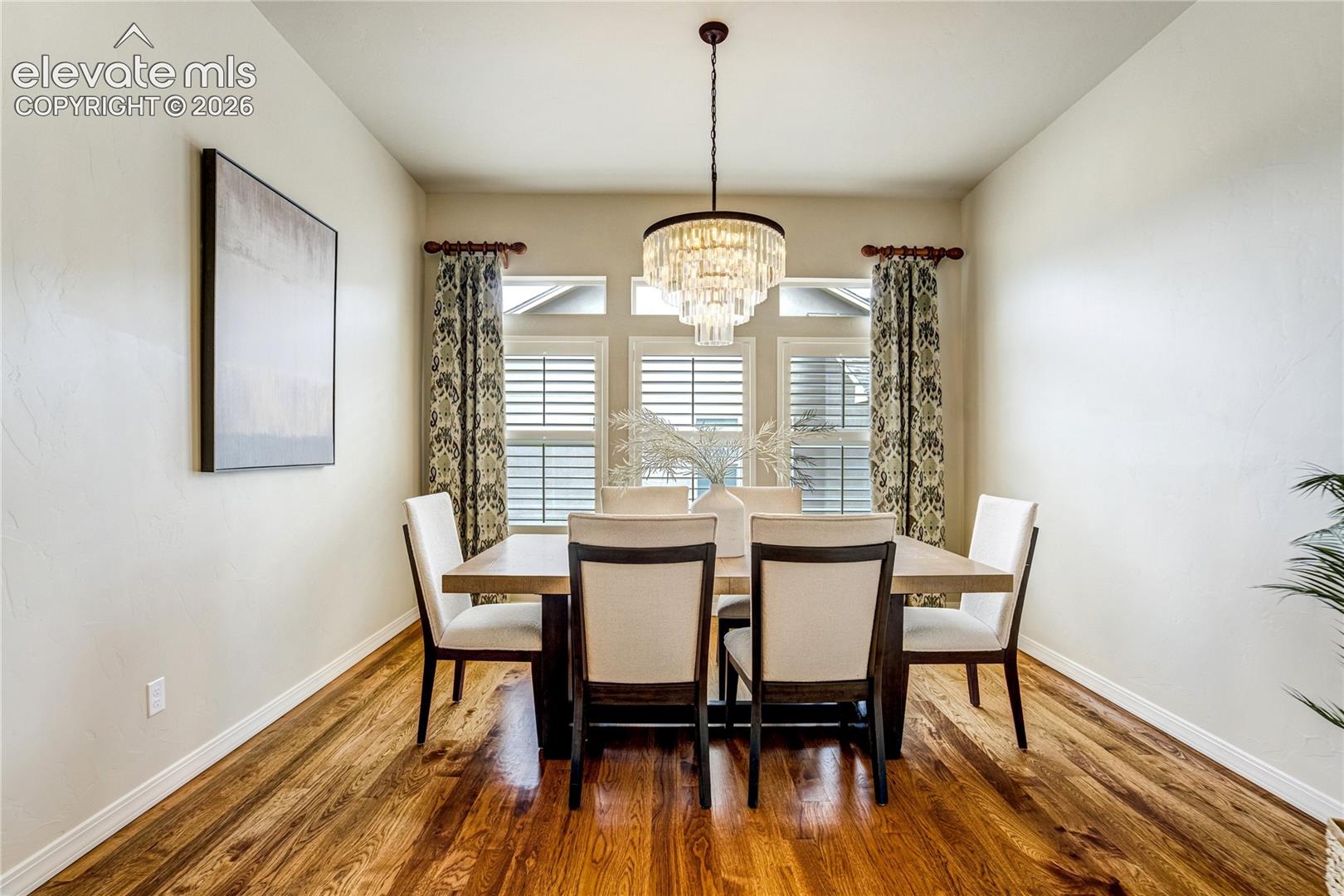 This elegant dining room features a central rectangular table surrounded by six upholstered chairs with dark wood frames. A tiered crystal chandelier hangs above the table, serving as a focal point, while patterned curtains frame the large windows that allow natural light to fill the space. The room is finished with warm-toned wood flooring and a large piece of abstract wall art, creating a sophisticated and inviting atmosphere.