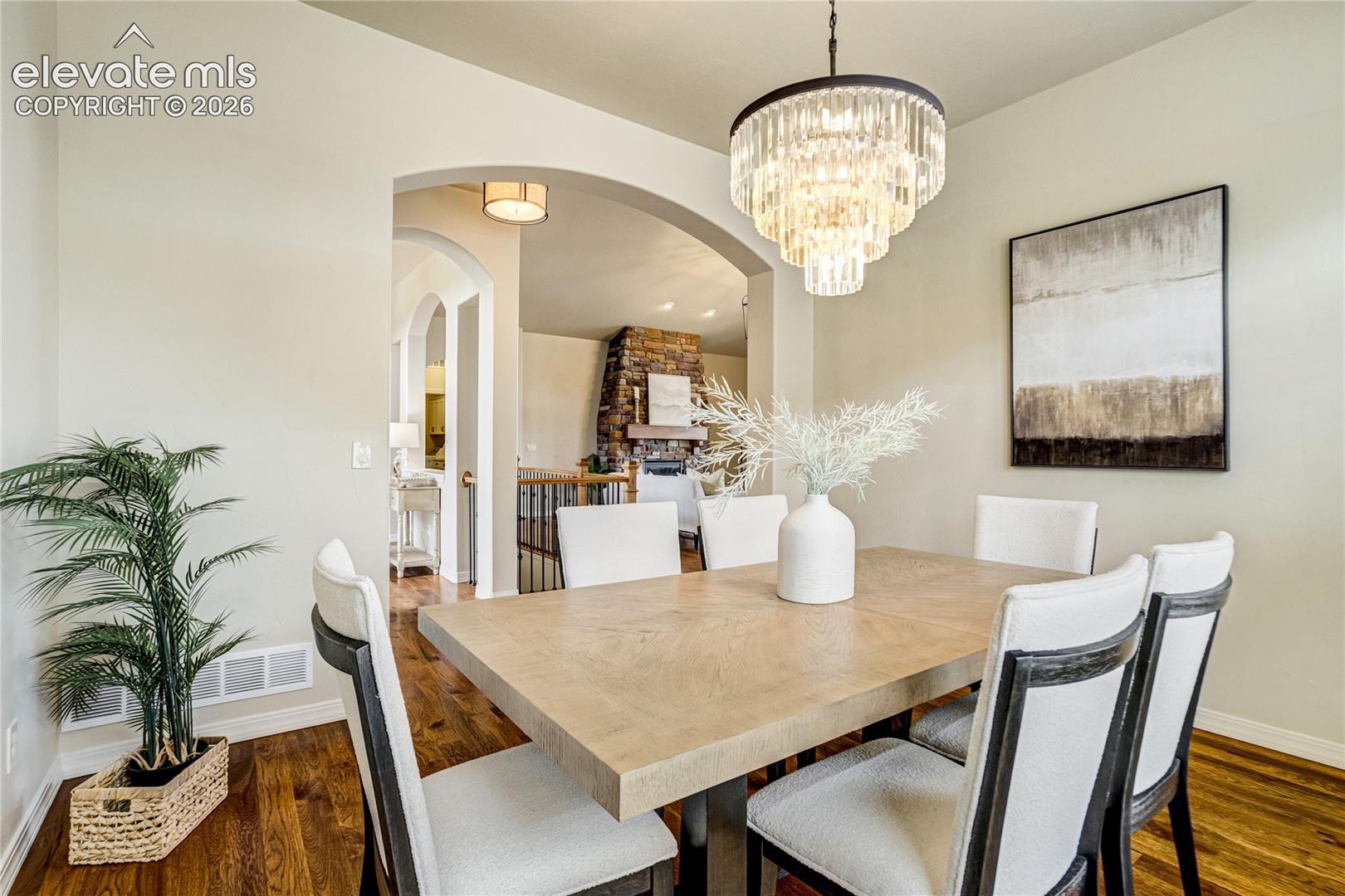 This elegant dining room features a light wood table surrounded by six upholstered chairs with dark frames, positioned beneath a prominent crystal chandelier. The space is decorated with a large abstract wall art piece and a potted plant in a woven basket, creating a sophisticated and inviting atmosphere. An arched opening provides a view into an adjacent living area, which showcases a stone-clad chimney breast and a wooden staircase.