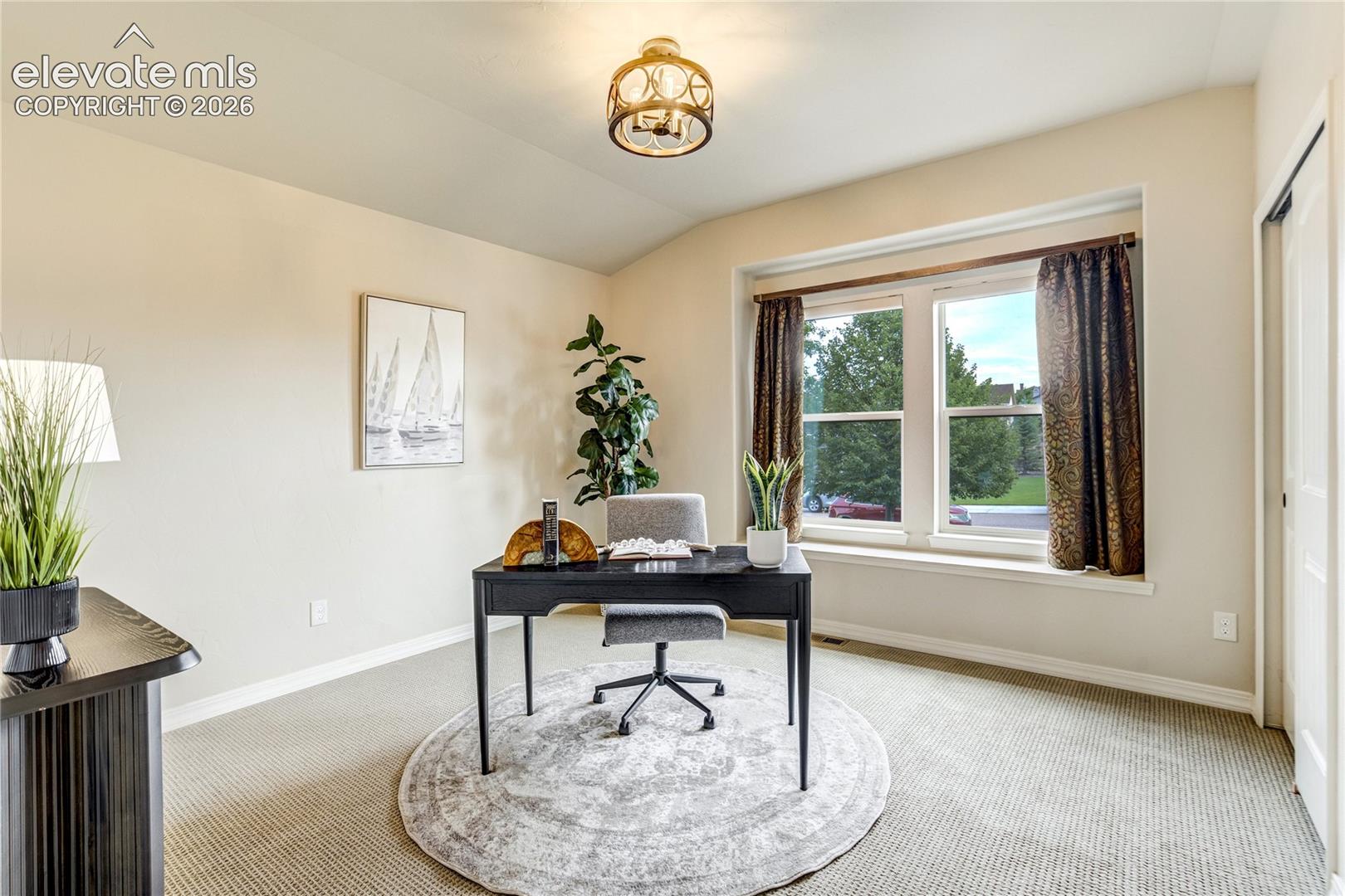 This bright and functional home office features a central black desk paired with a grey upholstered chair, set atop a circular patterned area rug. The room is accented by a tall potted plant, a sailboat-themed wall print, and a decorative light fixture, all illuminated by natural light from a large window with brown curtains. The neutral wall color and carpeted flooring create a professional yet inviting workspace.
