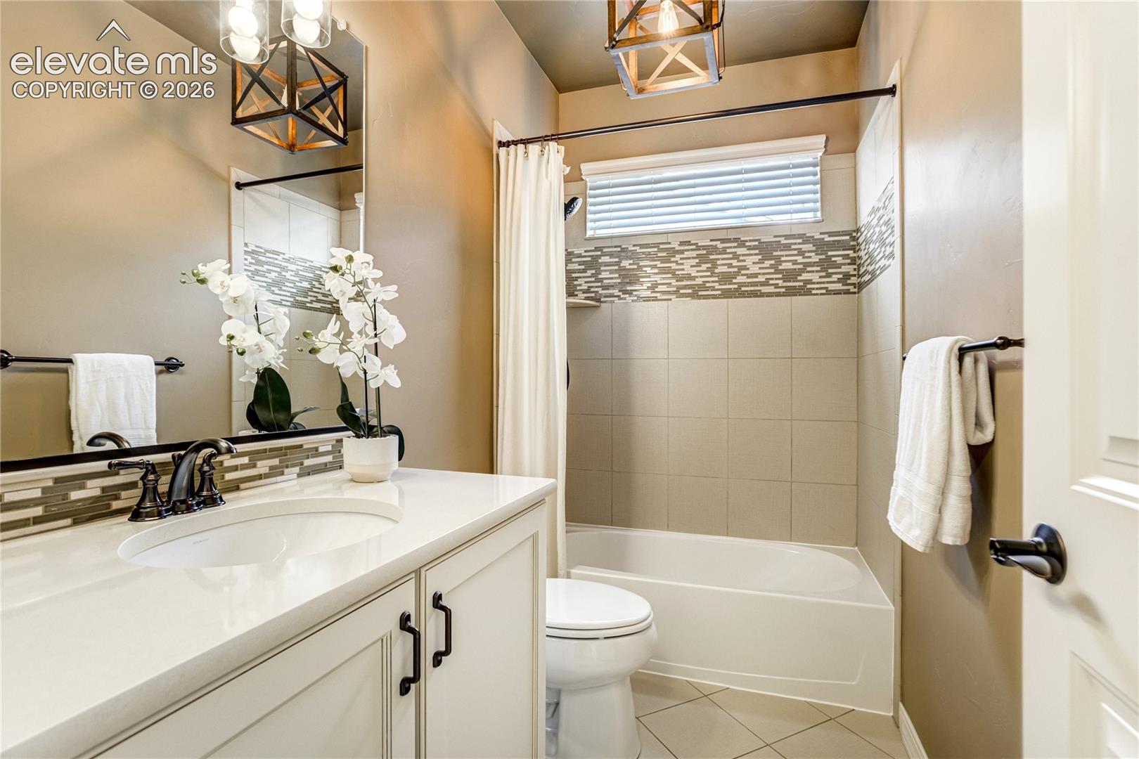 This compact, well-lit bathroom features a white vanity with a light-toned countertop and dark hardware, complemented by a large mirror and a decorative light fixture. A shower-tub combination is enclosed by a white curtain and includes a horizontal accent tile band that matches the backsplash at the vanity. The space is finished with neutral-toned wall paint and light-colored floor tiles, creating a clean and functional aesthetic.