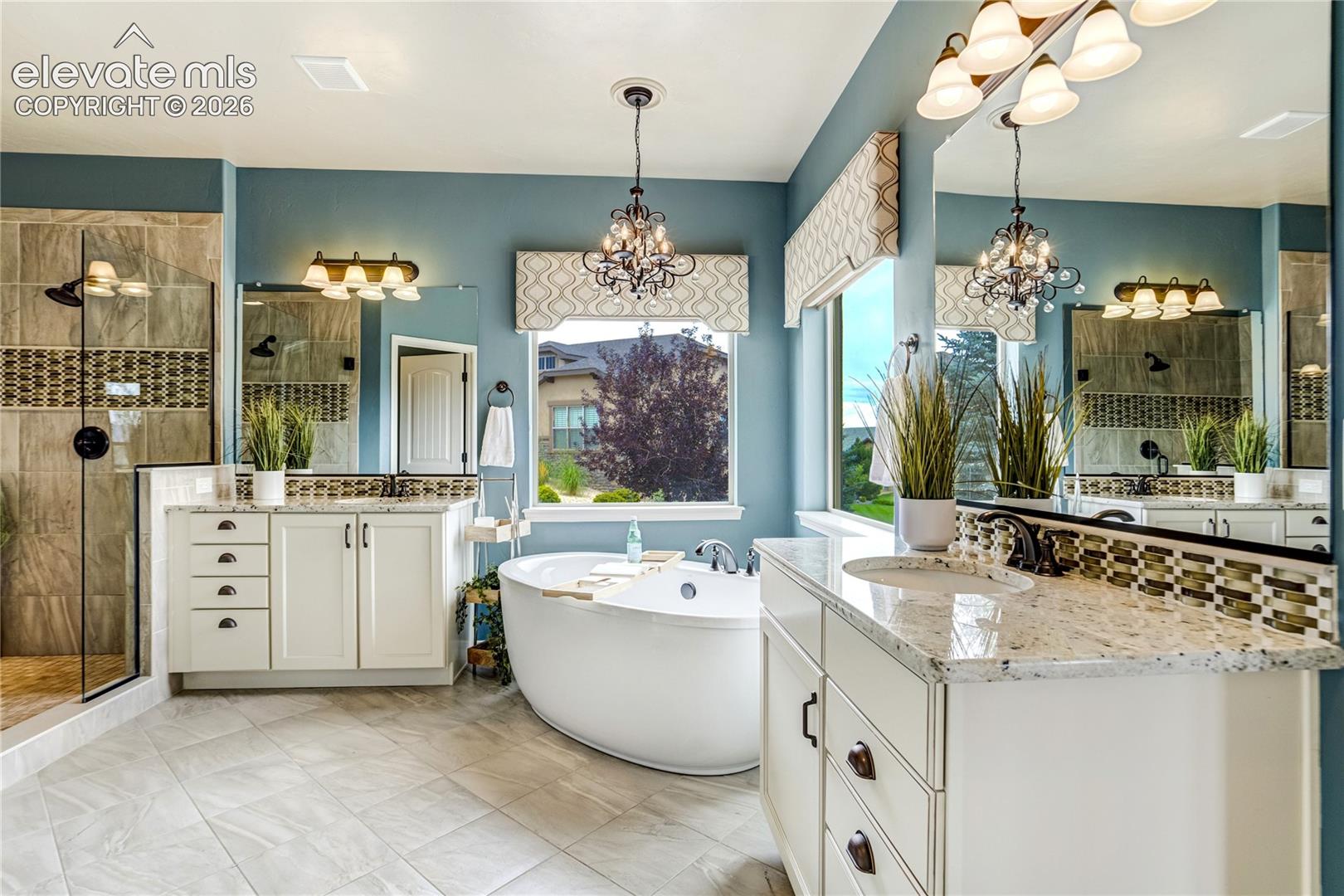 This spacious primary bathroom features a dual-vanity layout with white cabinetry, light-toned stone countertops, and large mirrors. A central freestanding soaking tub sits beneath a window with a decorative valance, while a glass-enclosed walk-in shower with tiled walls is positioned to the left. The room is accented by blue walls, elegant chandeliers, and neutral floor tiles, creating a bright and luxurious atmosphere.