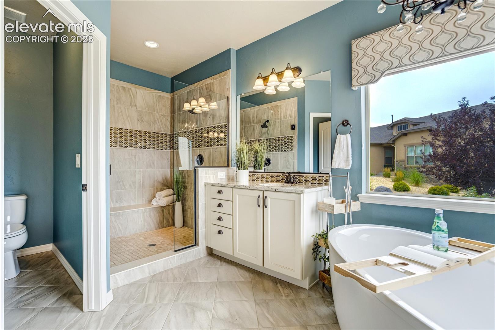 This primary bathroom features a spa-like atmosphere with a walk-in shower, a freestanding soaking tub, and a vanity with light-colored cabinetry. The space is accented by blue walls, patterned tile flooring, and a large window that provides natural light and a view of the neighborhood. The layout is functional and bright, highlighting a clean, modern aesthetic.