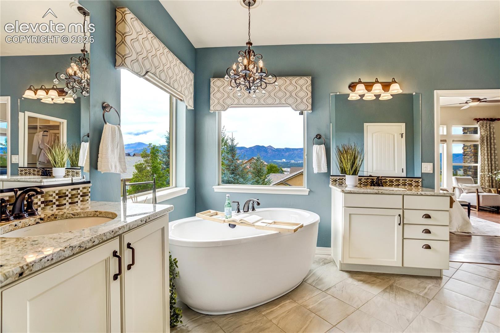 This primary bathroom features a central freestanding soaking tub positioned beneath a window with mountain views, accented by a decorative chandelier and patterned valances. The space includes dual vanities with light-toned stone countertops, dark hardware, and framed mirrors, all set against soft blue walls. The neutral tile flooring and clean, bright aesthetic create a serene and luxurious atmosphere.