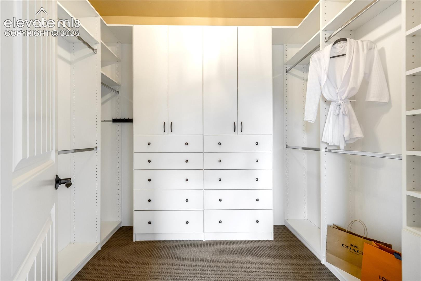 This organized walk-in closet features crisp white cabinetry with a symmetrical layout, including hanging rods on both sides and a central bank of drawers. The space is finished with dark, textured carpeting and clean, minimalist hardware on the drawers and cabinet doors. A white robe hangs on the right, and shopping bags are placed on the floor, suggesting a well-maintained and functional storage area.