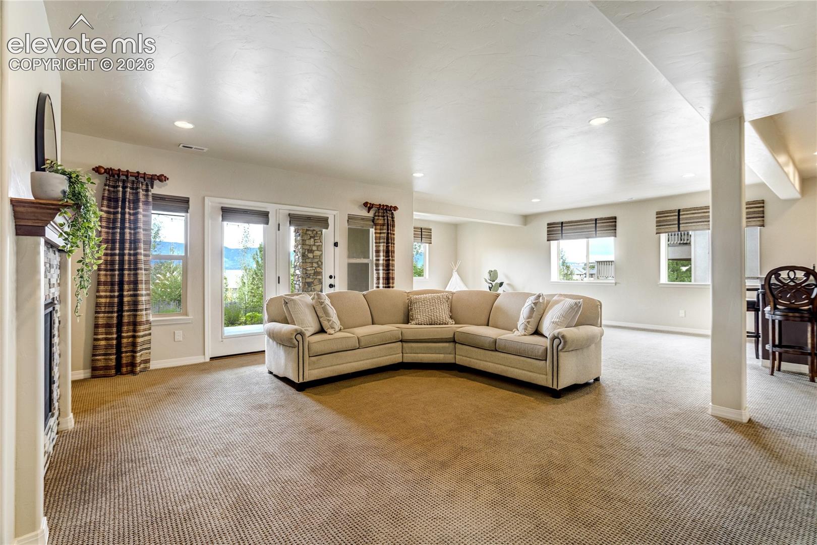 This spacious living room features a large, neutral-toned sectional sofa positioned as the central focal point of the open-concept area. To the left, a fireplace with a stone-clad chimney breast and a wooden mantel adds texture, while large windows and a glass door provide ample natural light and views of the outdoors. The room is finished with wall-to-wall carpeting and recessed lighting, creating a comfortable and inviting atmosphere.